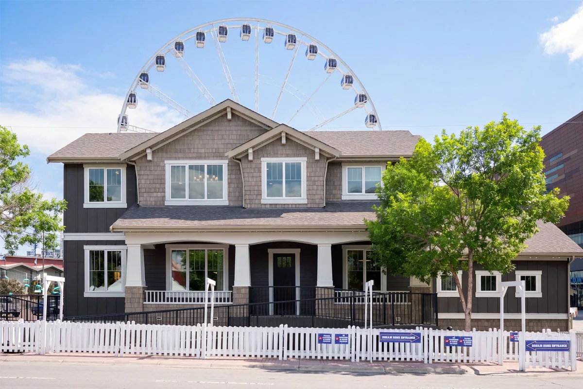 2026 Stampede Rotary Dream Home by Homes by Avi, a two-storey craftsman-style house with a covered front porch and white picket fence, set against the Calgary Stampede Ferris wheel